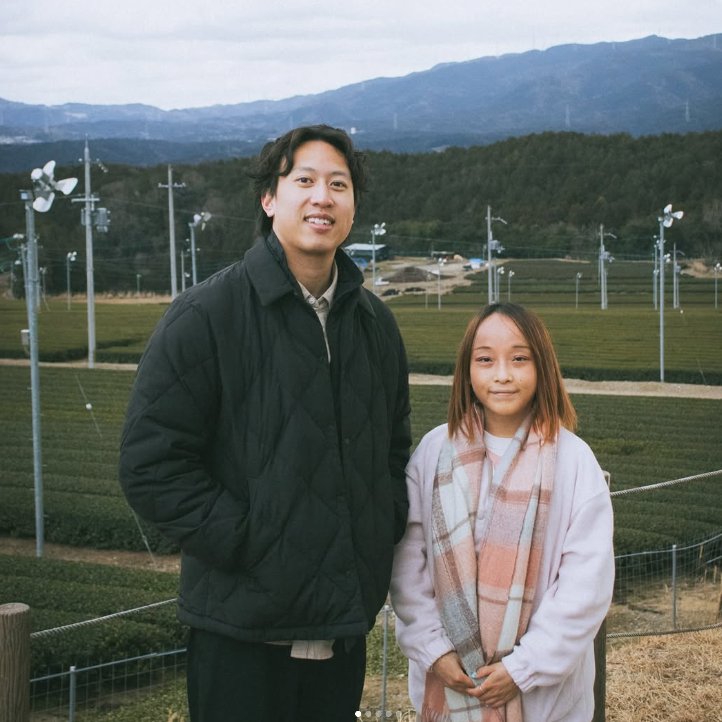 Principal owner of Gumo (Alex) standing beside matcha supplier (Shiori) outdoors at her farm with a scenic background of mountains and fields.